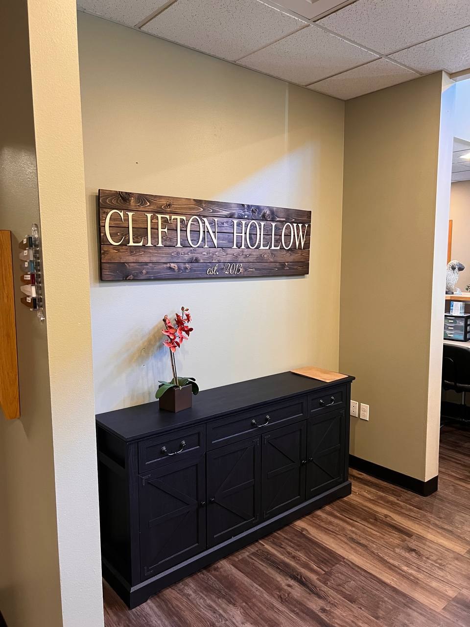 A carved wooden Clifton Hollow sign hanging above a cabinet with an orchid on it